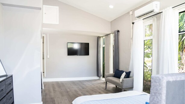 A bedroom with a bed, a TV mounted on the wall, a grey sofa, and a window with curtains.