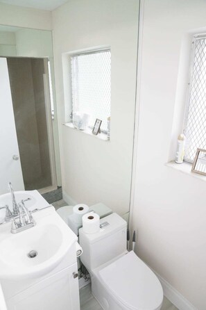 A modern bathroom with a white sink, toilet, and bidet. There is a shower area with a glass door, and a small window with a mesh screen above the toilet.