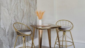 A corner of a room with a textured wall, a small round table with a white vase and pampas grass, and two gold-colored chairs with grey cushions. - Charlevoix Charm: Boujee Downtown Bungalow (Charlevoix)