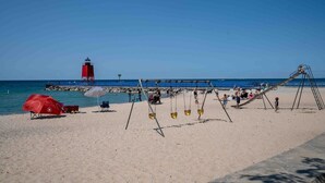 A beach with a swing set, a red umbrella, and a lighthouse in the background. - Charlevoix Charm: Boujee Downtown Bungalow (Charlevoix)