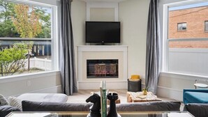 A living room with a fireplace, a television mounted above it, and a large window on one side. - Charlevoix Charm: Boujee Downtown Bungalow (Charlevoix)