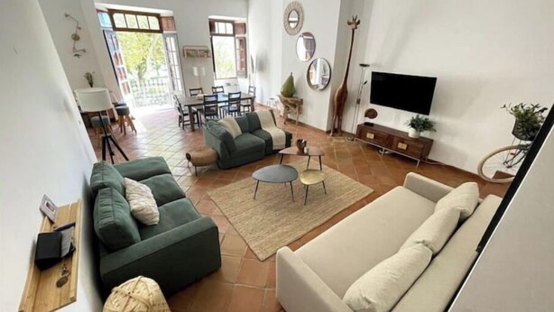 Un salón amplio con un sofá grande, un sofá más pequeño, una mesa de centro y un televisor. En el fondo, hay un comedor con una mesa y sillas.