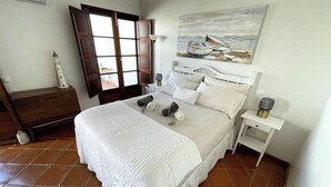 Una cama bien hecha con cubrecama y almohadas blancas, una mesita de noche con lámpara, un cuadro de barcos en la pared, y una ventana con vistas al exterior.