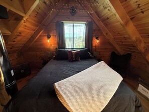 A cozy attic bedroom with a bed, two wall sconces, a window with curtains, and a small fan. - Relax at The Lincoln Log Cabin perched along the Jump River. (Sheldon)