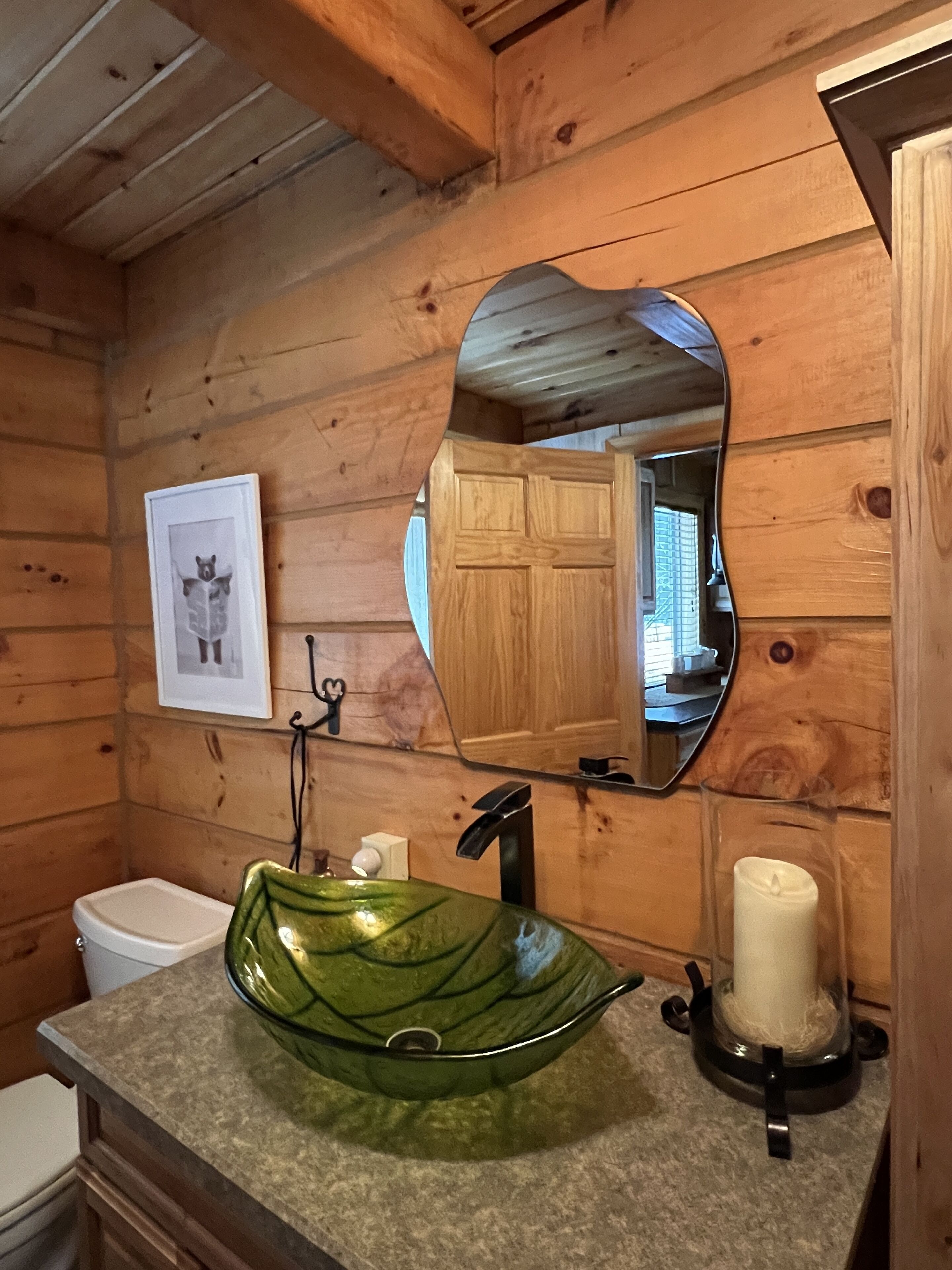 Un baño de cabaña de madera con un lavamanos de vidrio verde, un espejo y un cuadro en la pared.
