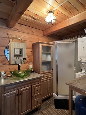 Interior de una cabaña de madera con un armario de madera, un lavamanos de vidrio verde, un espejo, una lámpara y un refrigerador de acero inoxidable.