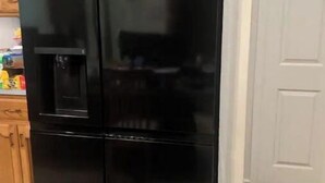 A black LG refrigerator in a kitchen with wooden cabinets and a white door in the background.