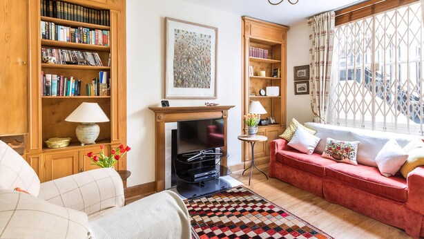 A cozy living room with a fireplace, a red sofa, a bookshelf, and a large window with curtains.