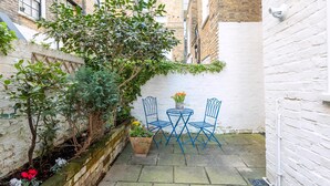 A narrow courtyard with a white brick wall, a small tree, and a table with blue chairs.