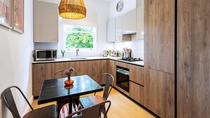 A modern kitchen with wooden cabinets, a dining table with chairs, a hanging light fixture, and a window with a view of greenery. - Stunning Earls Court Flat (London)