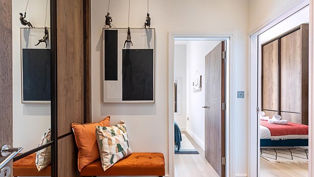 A modern hotel room with a wooden bench, a bed with a red blanket, and a hallway leading to another room. - Stunning Earls Court Flat (London)