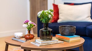 A round wooden coffee table with a clear glass vase containing white flowers, a book titled 'BURNOUT', and a ceramic bowl. A blue sofa with a white cushion is in the background. - Stunning Earls Court Flat (London)