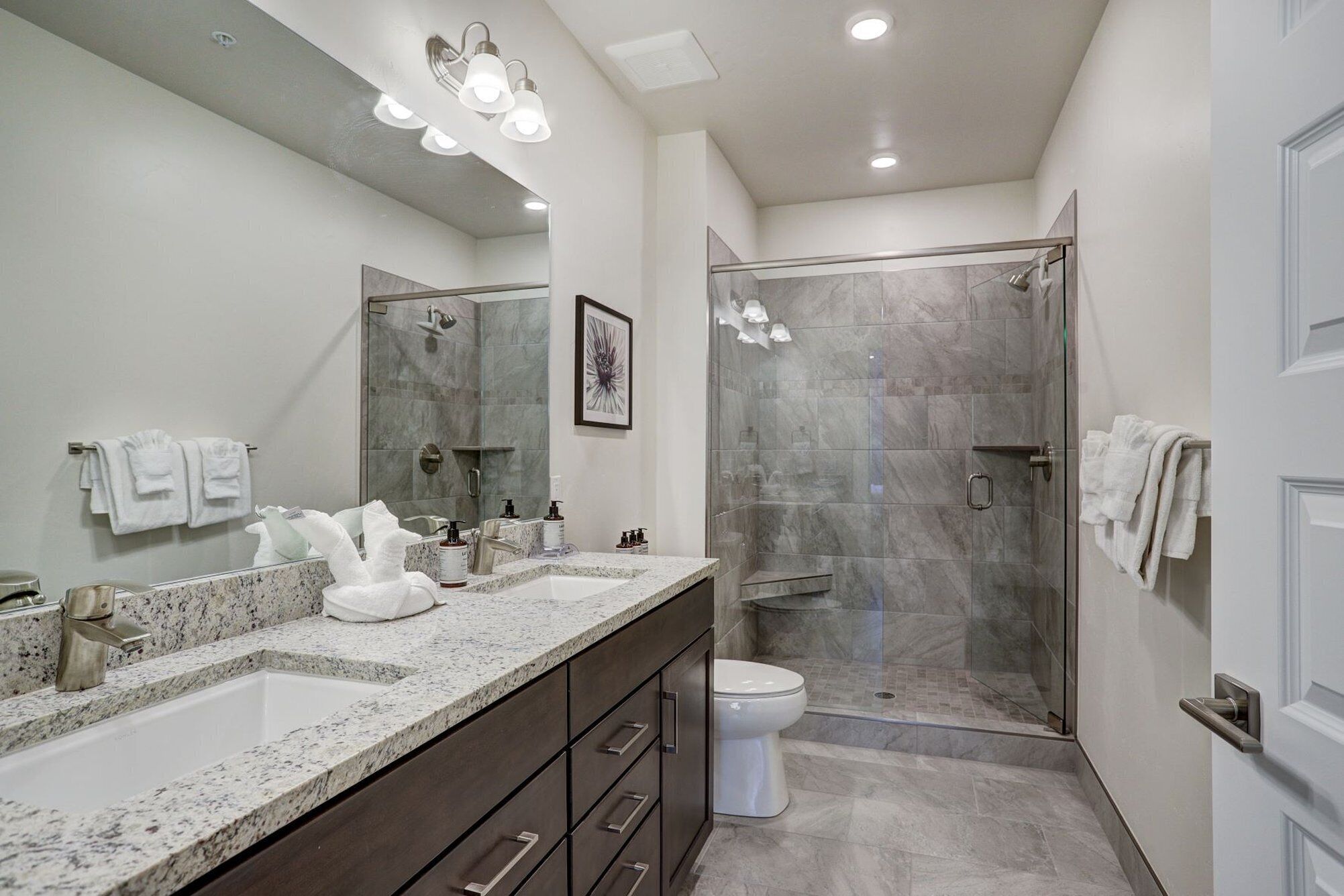 A modern bathroom with a large mirror, two sinks, a toilet, and a glass-enclosed shower.