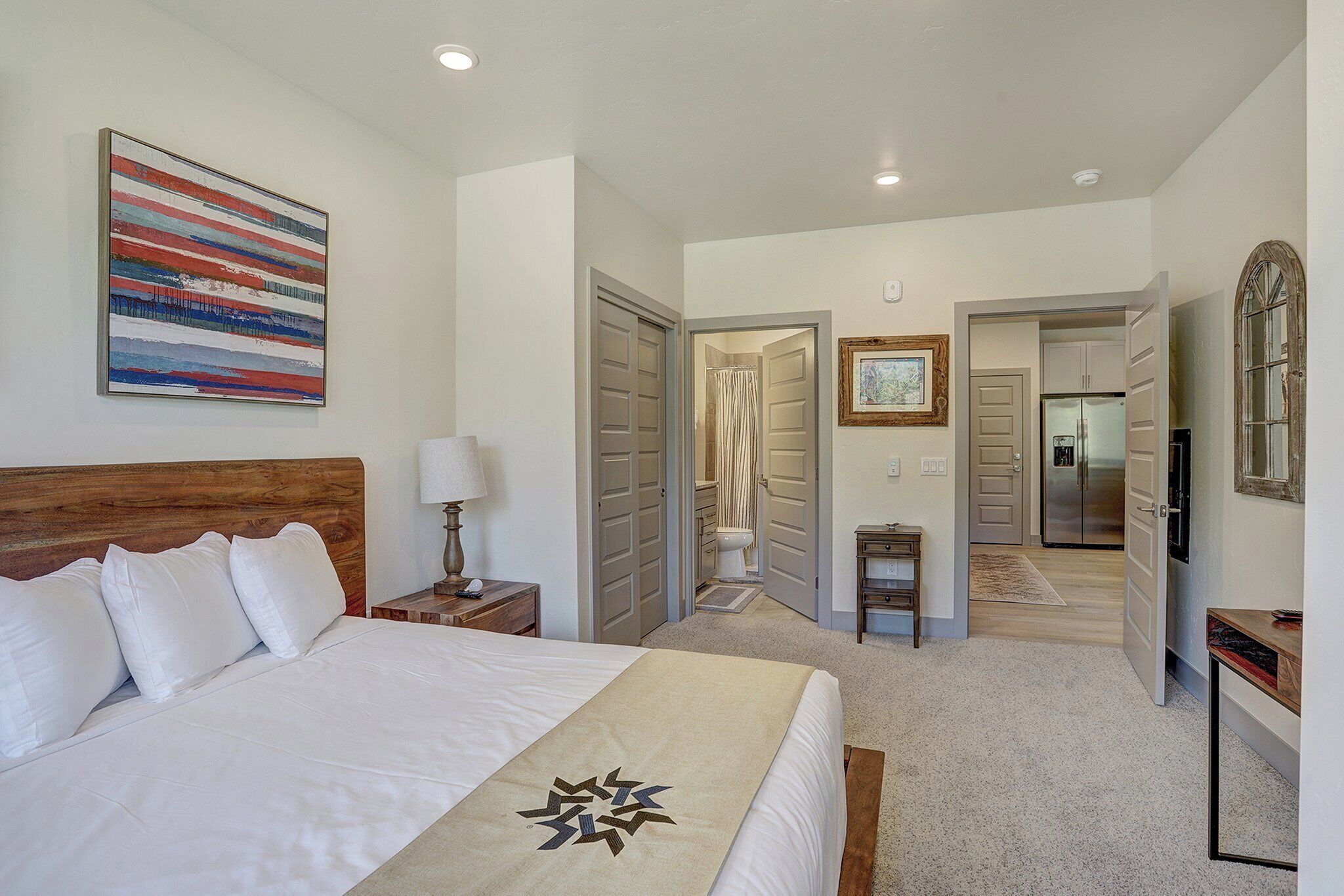 A hotel room with a large bed, a bedside table with a lamp, a painting on the wall, and a view of the bathroom and kitchen through an open door.