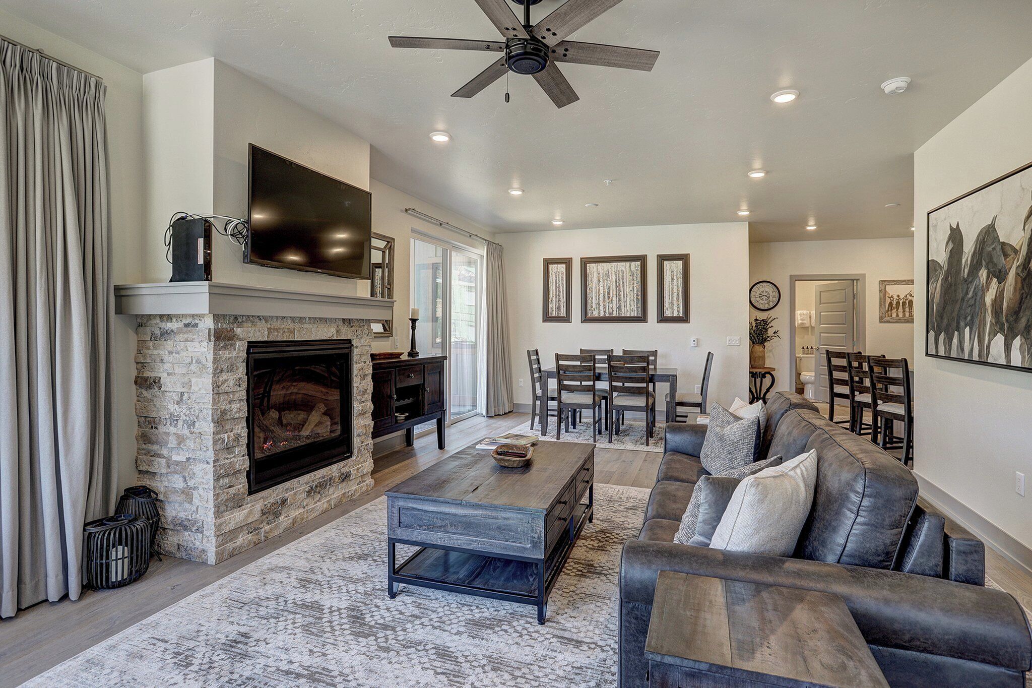 A spacious living room with a stone fireplace, a large flat-screen TV, a ceiling fan, and a dining area with a table and chairs.