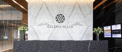 A reception area with a marble wall, a logo, and the text "GALERIA PLAZA".