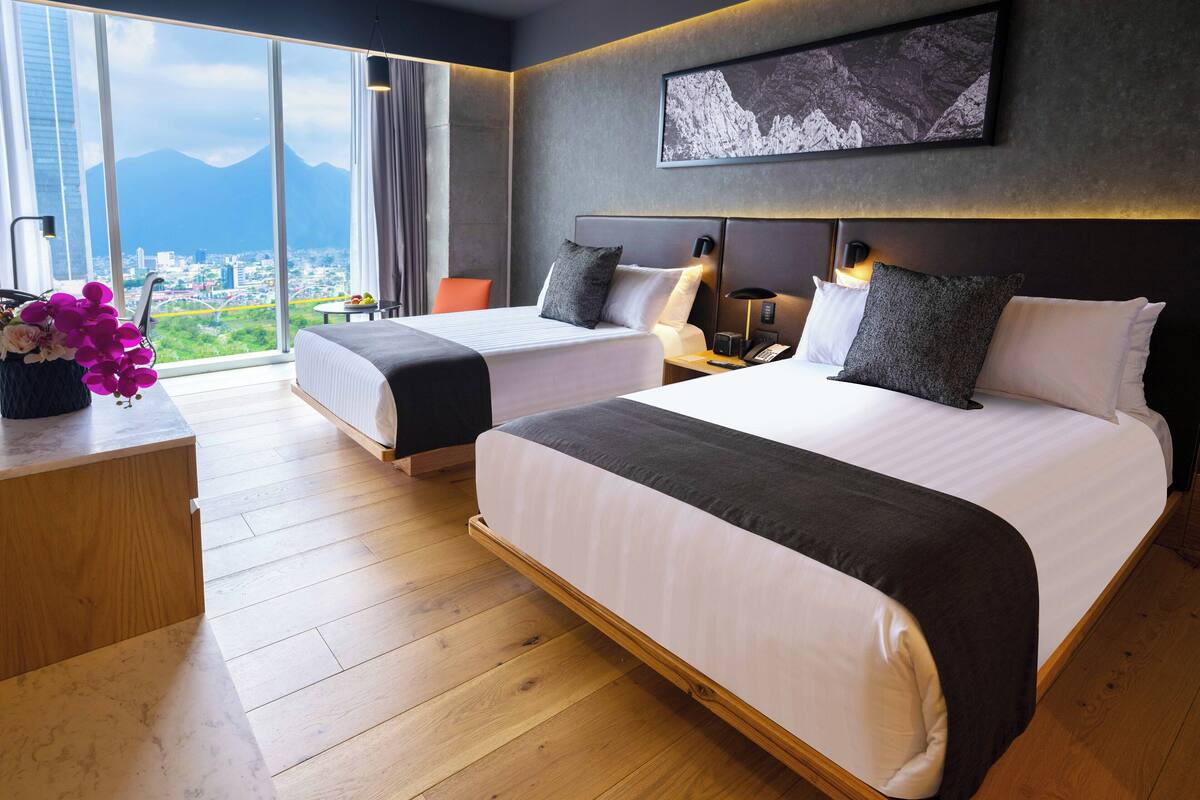 Une chambre d’hôtel avec deux lits, une grande fenêtre donnant sur la ville, une œuvre d’art accrochée au mur et un plancher en bois.