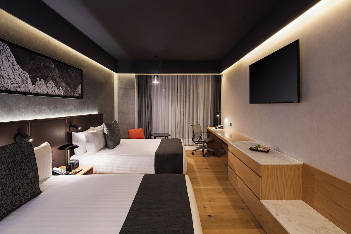 Une chambre d’hôtel moderne, équipée de deux lits, d’un bureau, d’une chaise, d’une télévision et ornée d’un tableau au mur.