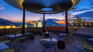 Un espacio al aire libre con mesas y sillas, un hogar de fuego y vistas a una ciudad y montañas al anochecer.