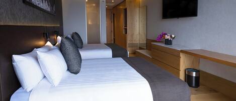 A modern hotel room with two beds, a flat-screen TV, a wooden desk, and a trash can.