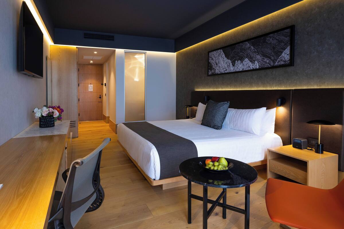 Une chambre d’hôtel moderne équipée d’un lit, d’un bureau, d’une chaise et d’une petite table sur laquelle se trouvent des fruits.
