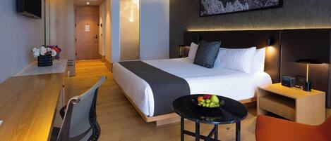 Una habitación de hotel moderna con una cama, escritorio, silla y una mesita con fruta.