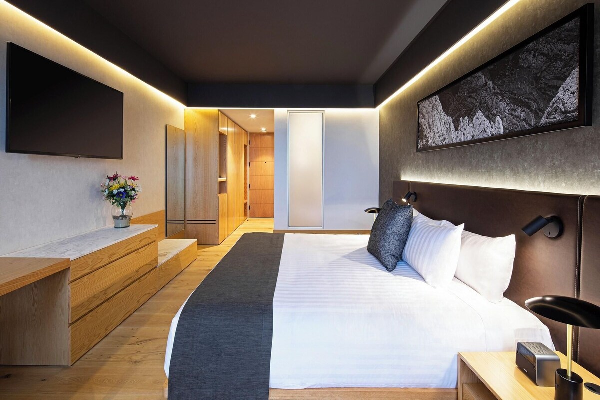 Une chambre moderne avec un grand lit, des tables de chevet, un téléviseur à écran plat, une œuvre d’art accrochée au mur et un banc en bois.