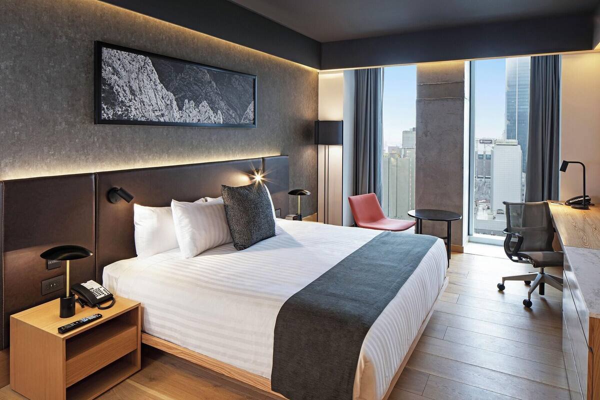 Une chambre d’hôtel moderne, dotée d’un grand lit, d’un bureau, d’une chaise, d’une lampe de bureau, d’un téléphone sur la table de chevet et offrant une vue sur la ville par la fenêtre.