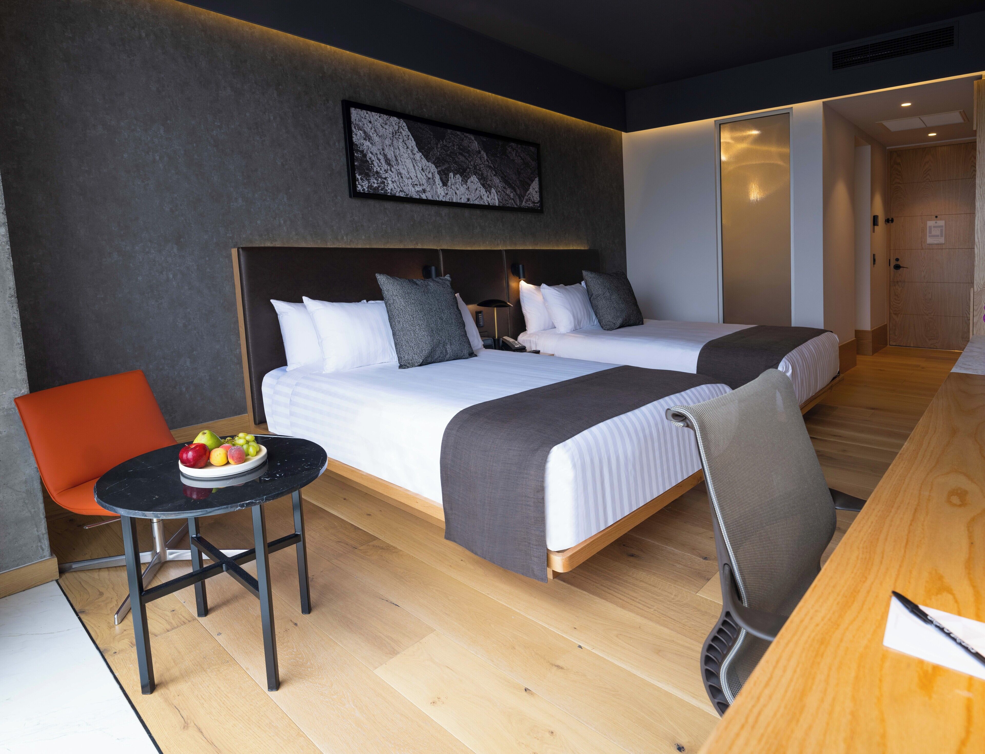 A hotel room with two beds, a dining table, a chair, and a fruit bowl.