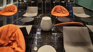 Zona de bar moderna con suelo de mármol negro, diversas opciones de asiento con sillas naranja y grises, y una unidad de estanterías central que exhibe varios objetos.