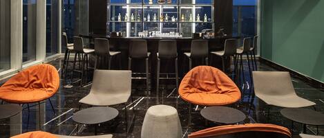 Un espace bar moderne avec un sol en marbre noir, de nombreux types de sièges, notamment des chaises orange et grises, et une étagère centrale où sont exposés divers objets.