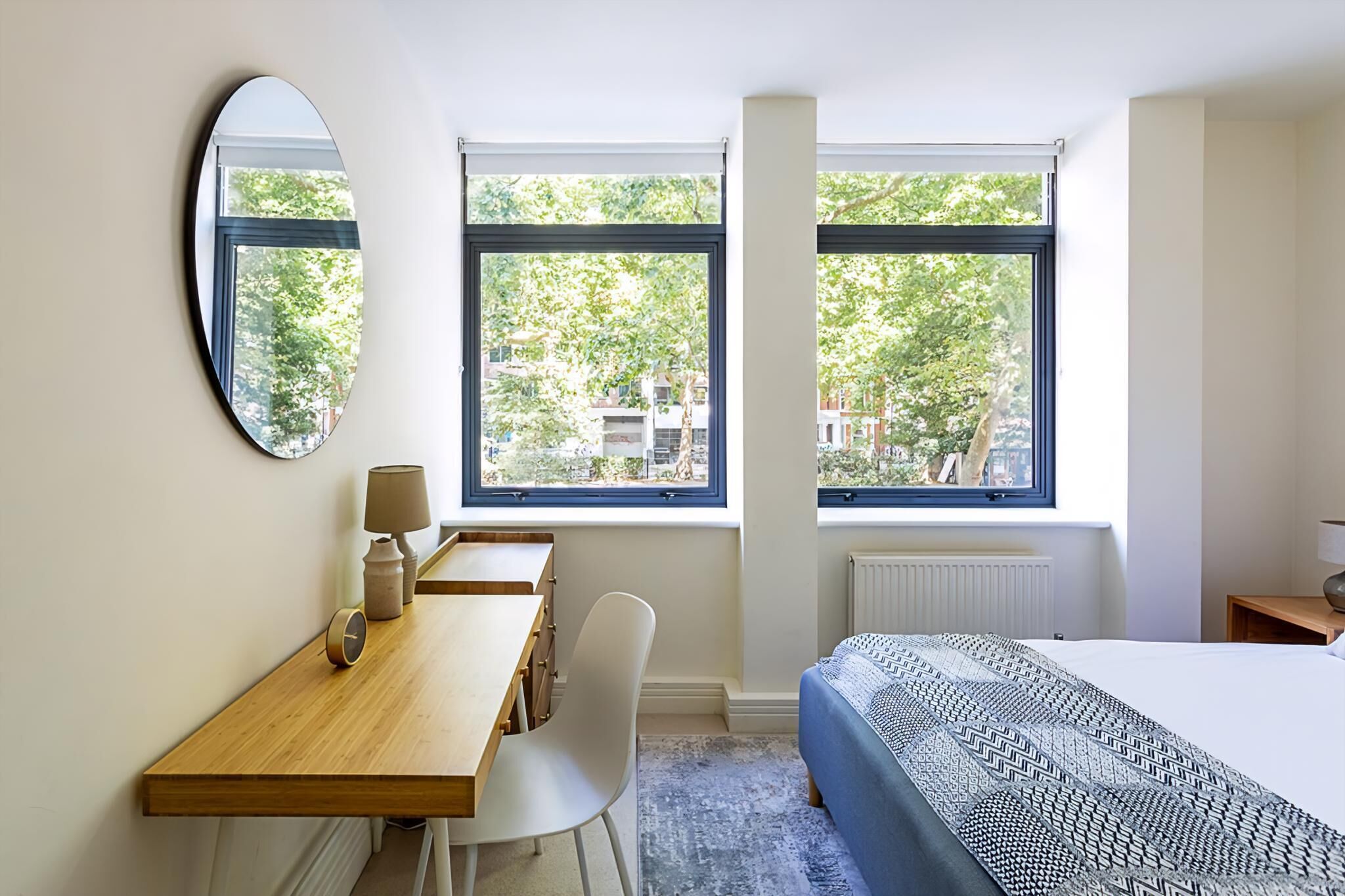 A bedroom with a bed, a desk with a chair, a lamp, a mirror, and two windows with a view of trees and buildings.