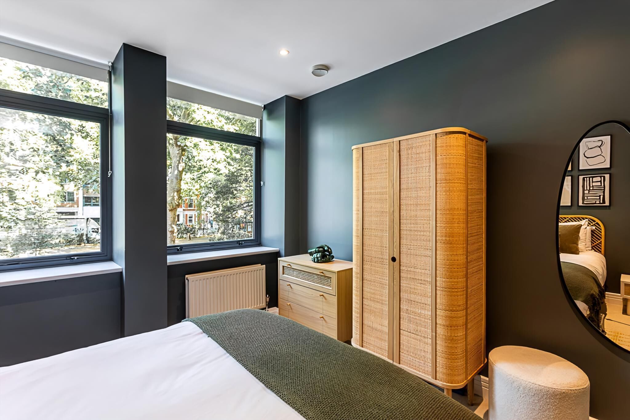 Une chambre à coucher comprenant un lit, une commode et une armoire, de grandes fenêtres et une vue sur les arbres et les bâtiments environnants.