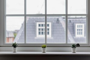 Ein Fenster mit drei Blumentöpfen auf einem Sims, mit Blick auf ein Gebäude mit vielen Fenstern.