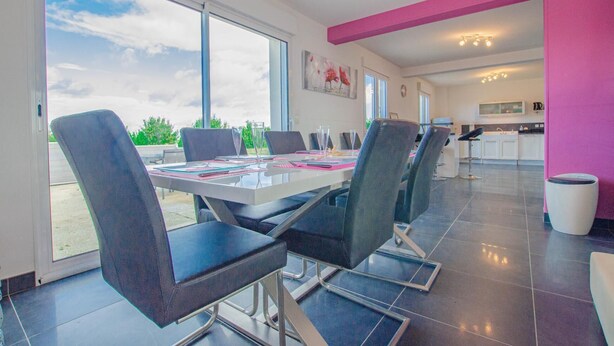 Un espace repas moderne avec une table en verre et des chaises noires, de grandes fenêtres et une cuisine en arri ère-plan.