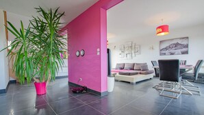 Un salon moderne avec un mur d’accent rose, une grande plante en pot, un canapé et un coin repas comprenant des chaises et une table.