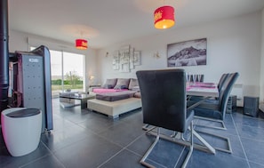 Ein modernes Wohnzimmer mit einer Couch, einem Esstisch mit Stühlen, einem großen Fenster und einer roten Pendelleuchte.