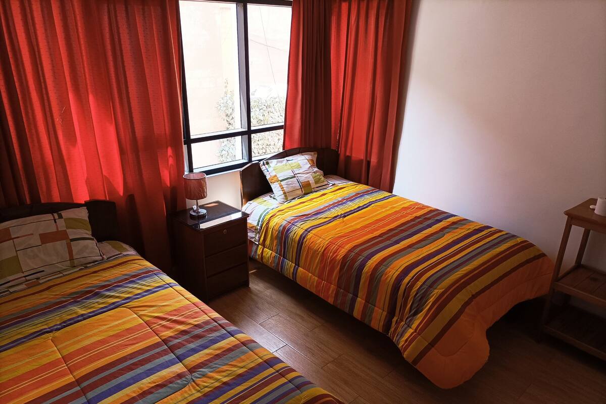 A bedroom with two beds, a nightstand, a window with red curtains, and a wooden floor.