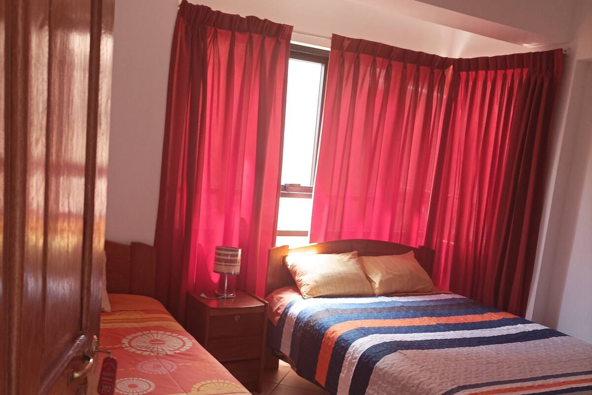 A bedroom with two beds, a nightstand, a window with curtains, and a door.