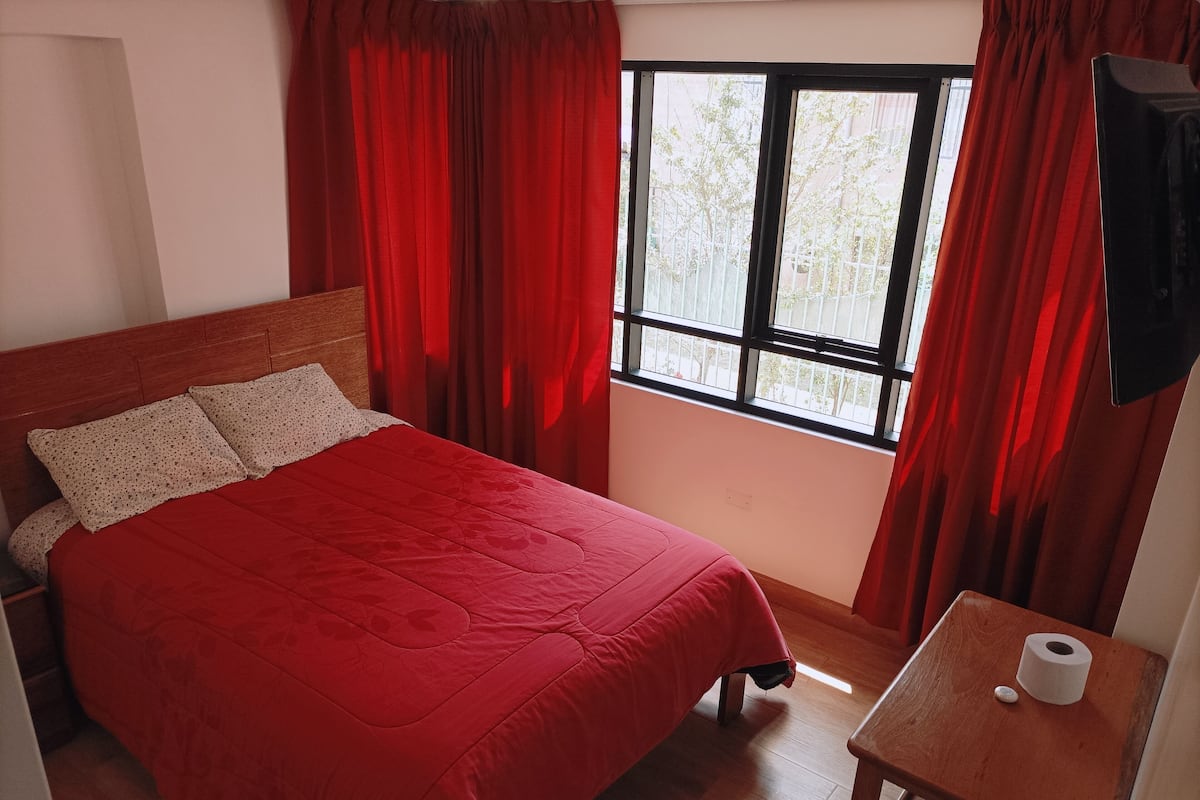 A bedroom with a bed, red curtains, a window, and a small table with a roll of toilet paper.