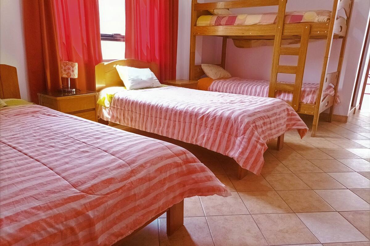 A room with two bunk beds, a bedside table with a lamp, and a window with red curtains.