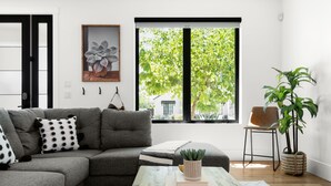 Una sala de estar moderna con un sofá modular gris, una mesa de centro de madera con libros y una planta en maceta, un ventanal con vista a zonas verdes, y una obra de arte colgada en la pared.