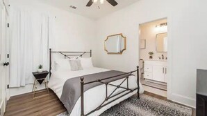 Un dormitorio con una cama, un ventilador de techo, una mesita de noche con una planta y un baño visible a través de una puerta abierta.