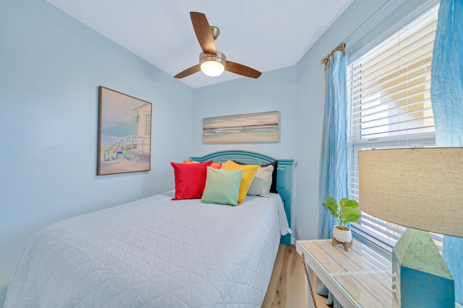 Quarto com cama, ventoinha no teto, janela com cortinas e duas pinturas na parede.