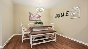 A dining area with a wooden table, white chairs, and a bench. There is a chandelier above the table and a floral painting on the wall. The word "HOME" is displayed on the wall. - Minutes from LSU, SU, & Downtown Baton Rouge! (Brusly)