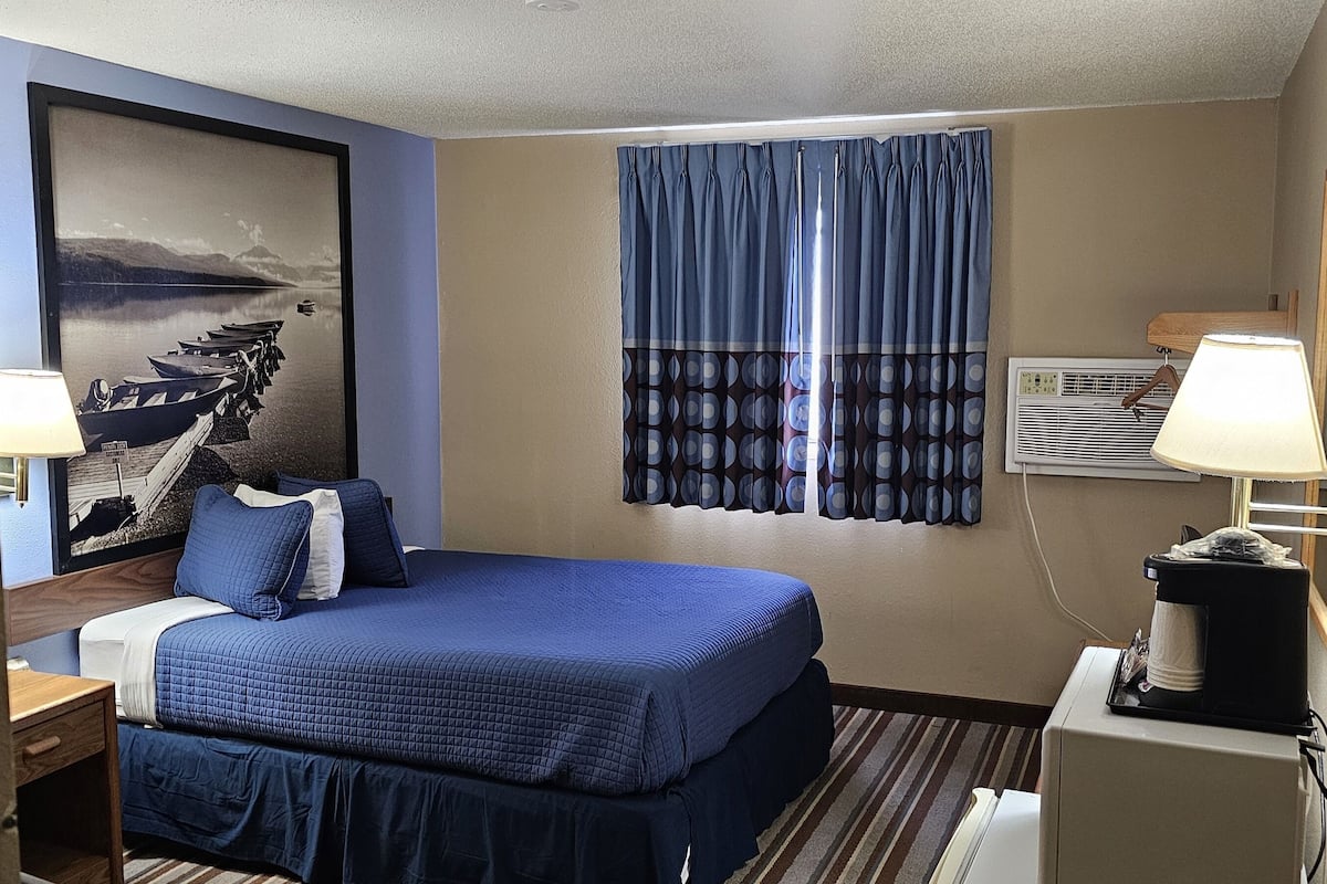 Quarto de hotel com cama, escrivaninha, luminária, ar condicionado e um quadro na parede.