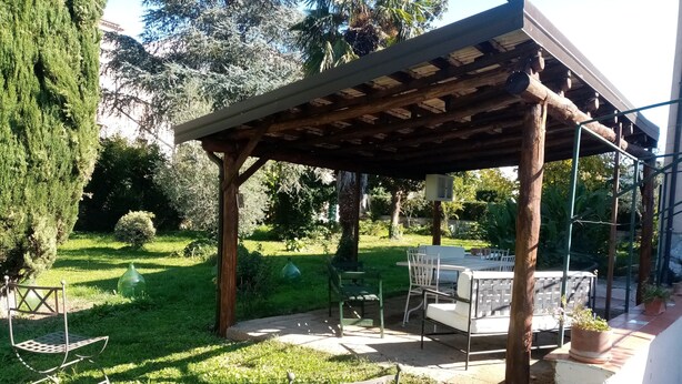 A covered outdoor seating area with a table and chairs, surrounded by greenery and trees.