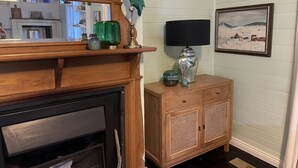 Een woonkamer met een open haard, houten mantel en een klein kastje met decoratieve objecten.