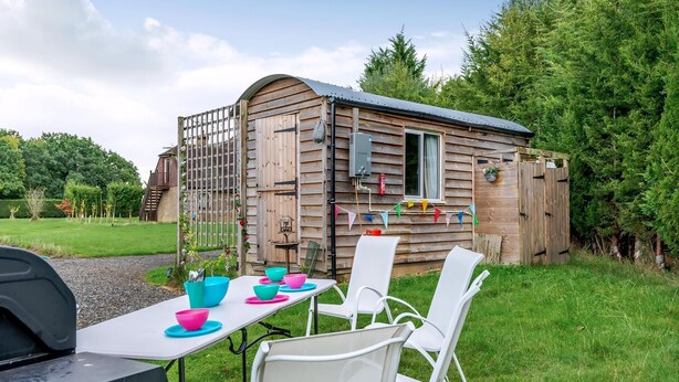 Een houten hutje met een klein terras, een tafel met stoelen en een barbecue.