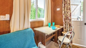Een knus vertrek met een houten wand, een raam met gordijnen, een blauwe bank, een kleine tafel met bekers en een kom, en een stoel.